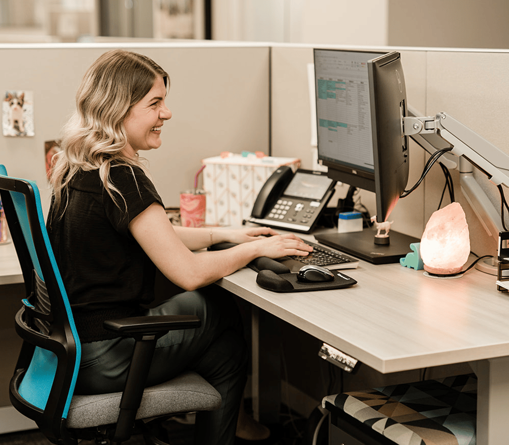 a woman sitting at a desk with a laptop and smiling at the camera